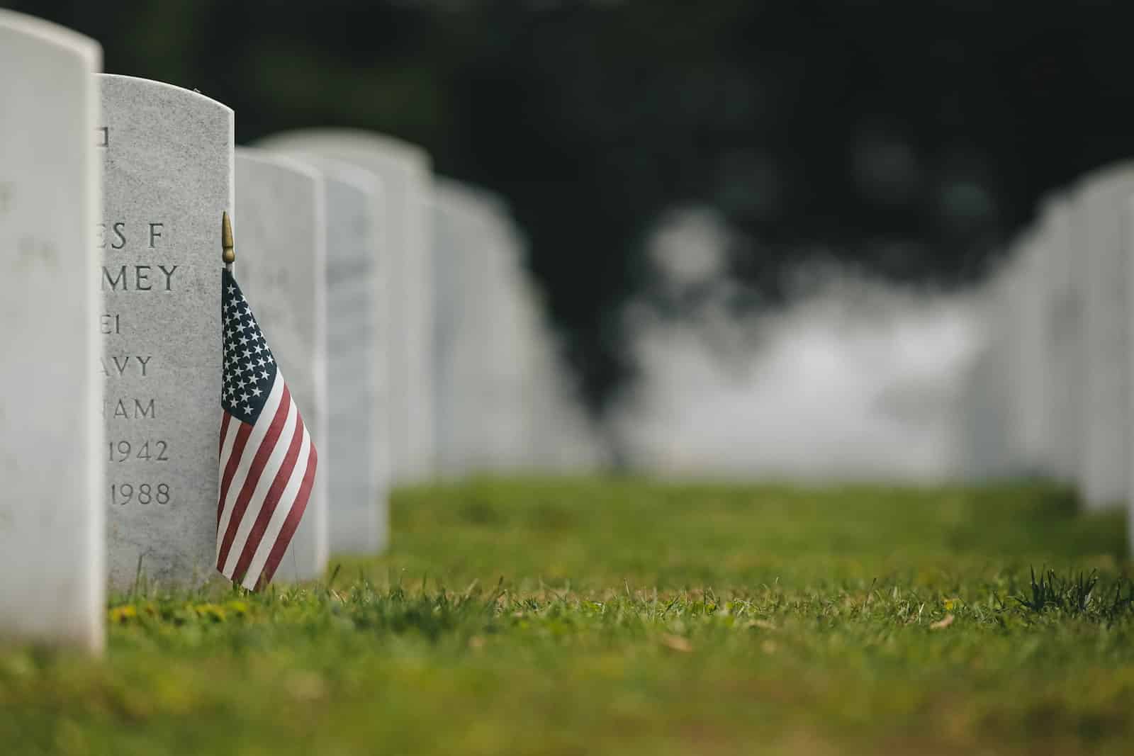 A small American flag against a white marble headstone in a national cemetery on a half-staff observance day